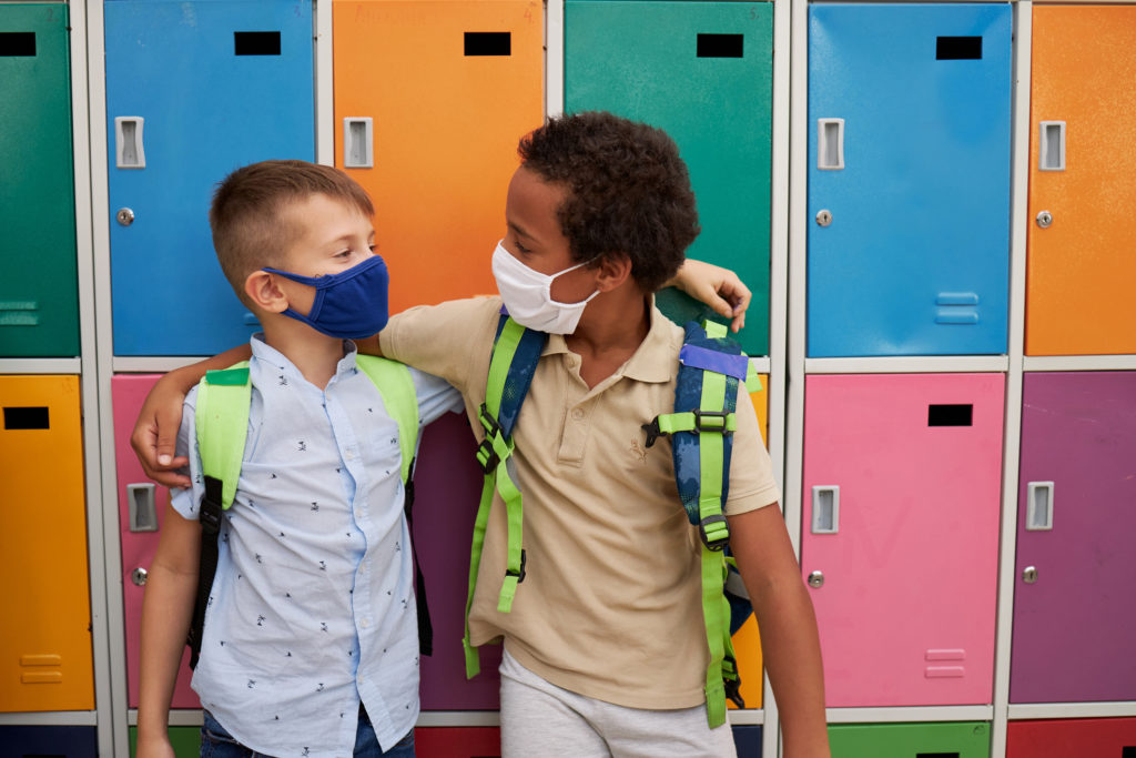 Cheerful multiracial male elementary students hugging in front of colorful lockers while wearing protective face mask during COVID-19 pandemic