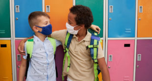 Cheerful multiracial male elementary students hugging in front of colorful lockers while wearing protective face mask during COVID-19 pandemic