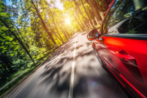 A car goes fast. The car is red. It is on a road in the forest. The sun shines brightly. 