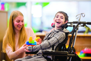 A caregiver is helping a young boy with a physical disability play with plastic block toys.