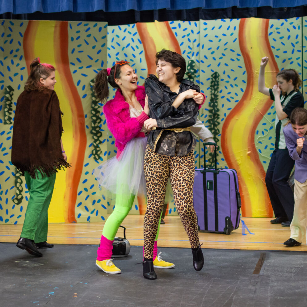 Two teens in bright costumes laugh on a stage. 