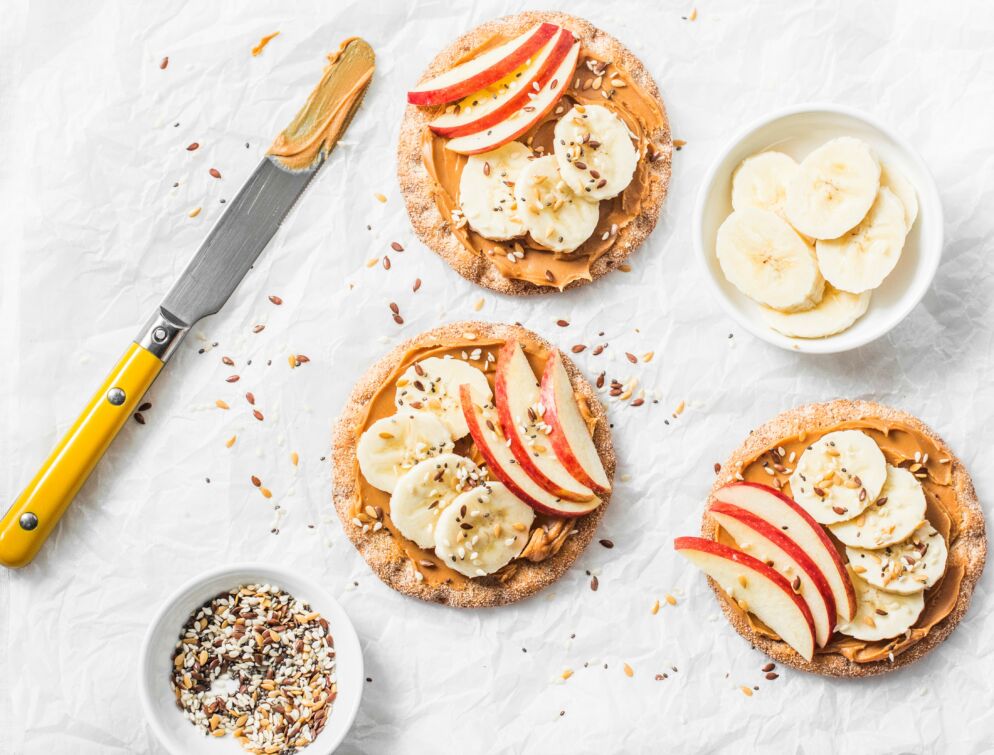 A butter knife smeared with peanut butter rests on a countertop next to some crackers which are topped with peanut butter, banana slices, apple slices and nuts. 