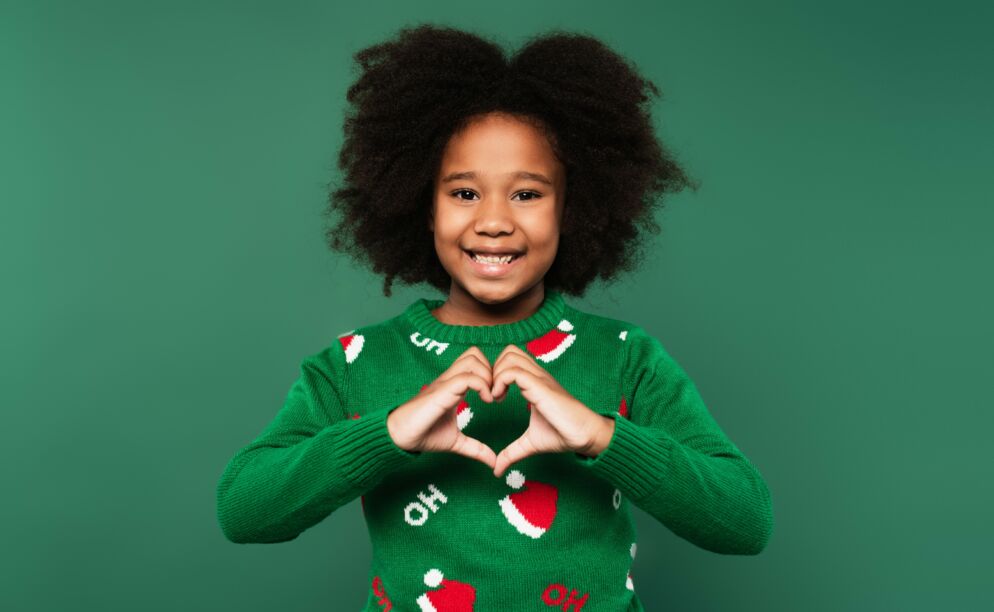 A young girl makes a heart shape with her hands. She is wearing a green sweater with little Santa hat designs.