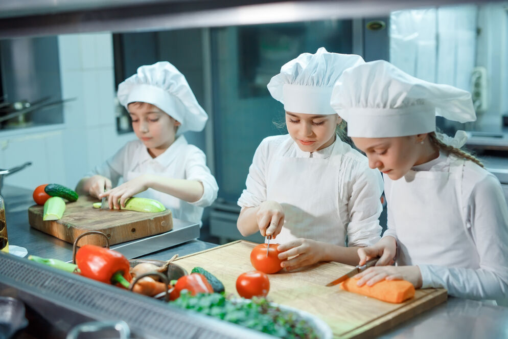 Children in white chef's clothing chop vegetables. 