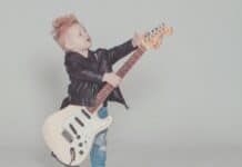 A young boy with a blond hair in a mohawk style wearing a leather jacker holds an electric guitar.