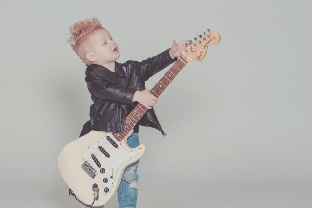 A young boy with a blond hair in a mohawk style wearing a leather jacker holds an electric guitar.