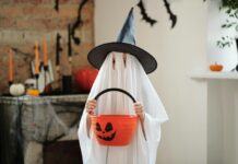 A child dressed in a ghost costume and wearing a witch hat holds up a bucket of Halloween candy