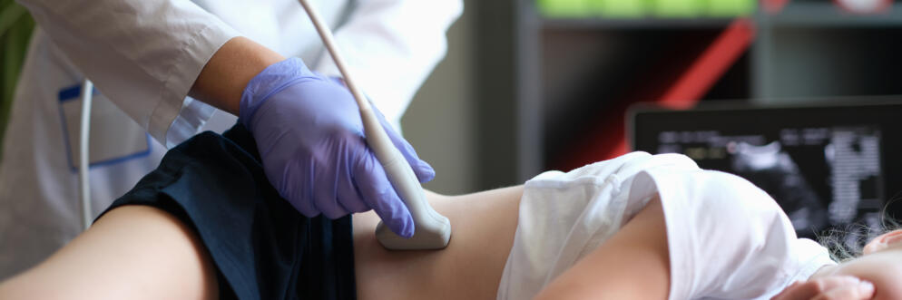 Doctor conducting ultrasound examination of kidneys to child in clinic. The child is laying on their side as a doctor wearing gloves and a white coat places the instrument on her skin.