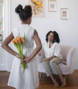 A girl holds flowers behind her back to give her mother