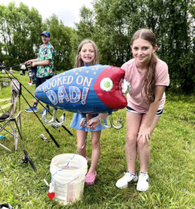 Two girls, one holds up a large fish-shaped balloon that says "Hooked on Dad!"