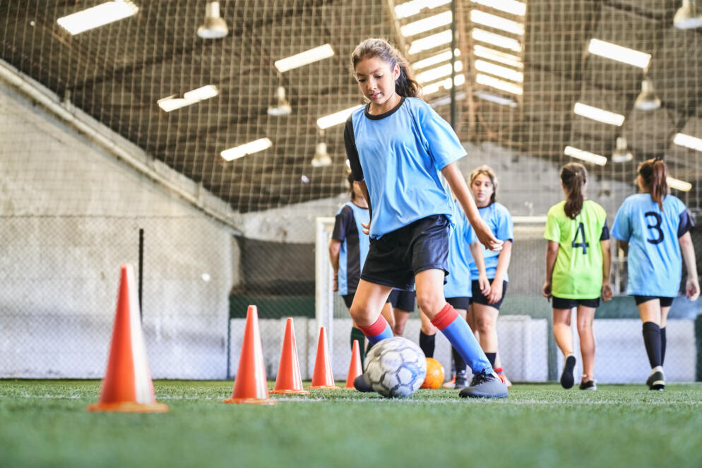 Young girl leading ball between cones on sports court. Female player in blue jersey practicing dribbling skill at court.